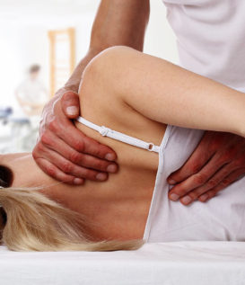 This image shows a woman laying on a table. There are hands on her shoulder blade, part of full body chiropractic adjustments.